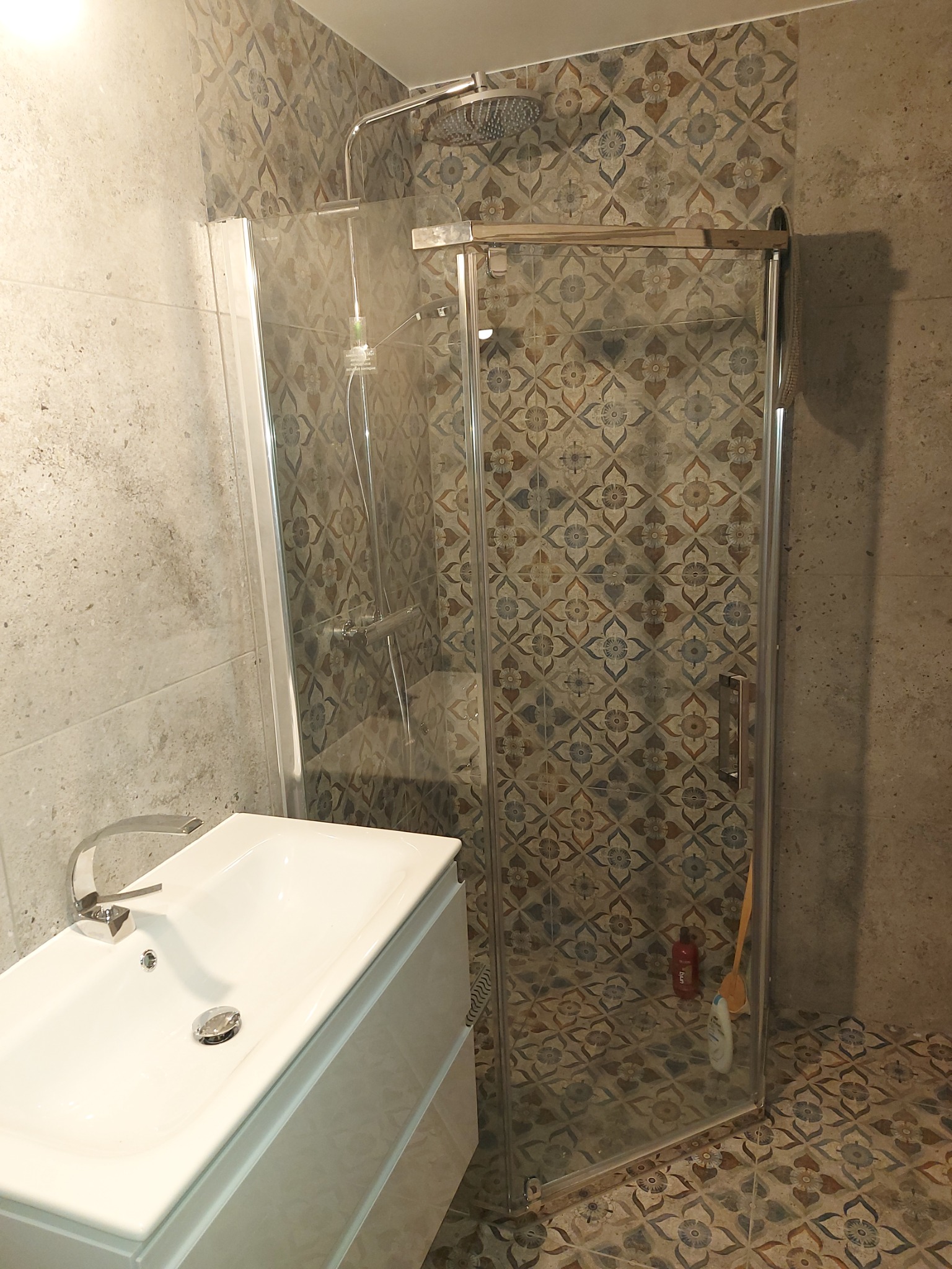 Nowoczesna łazienka z kabiną prysznicową ze szklanymi drzwiami, umywalką z szafką w kolorze białym i mozaiką na ścianach i podłodze w odcieniach szarości, brązu i niebieskiego.