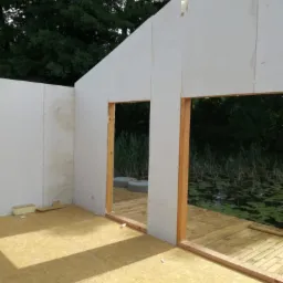 Wnętrze szkieletowej konstrukcji budynku w trakcie budowy, widoczne płyty OSB na podłodze i ścianach, drewniane ramy okienne, z zewnątrz widok na zarośla i staw.