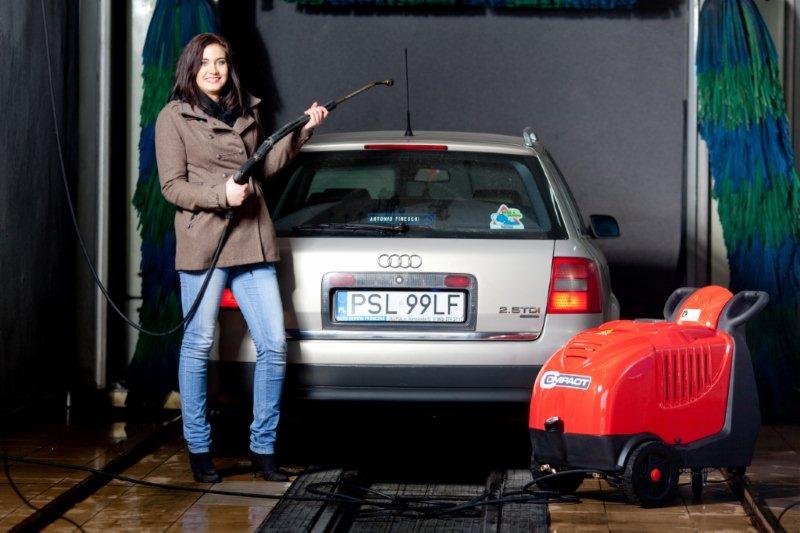 Kobieta uśmiechnięta trzyma lancę myjki ciśnieniowej przed srebrnym Audi kombi w myjni automatycznej; widoczna czerwona myjka ciśnieniowa Compact.