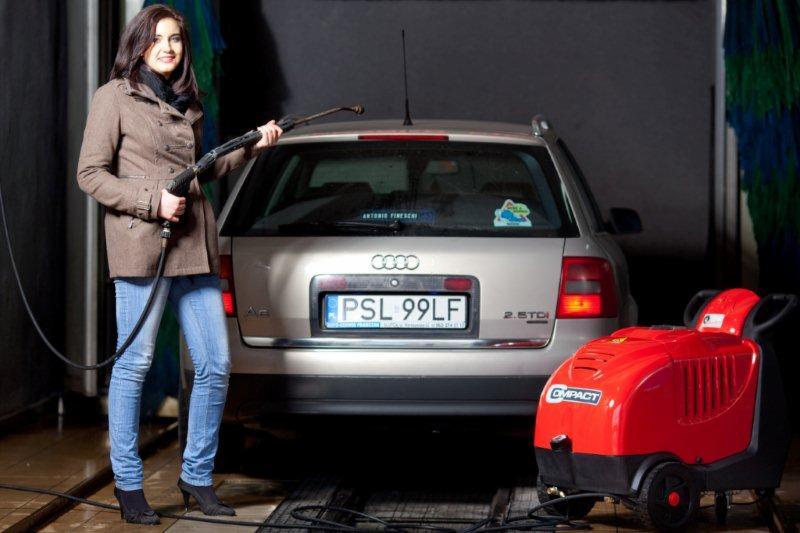 Kobieta w jeansach i brązowym płaszczu trzyma lancę myjki ciśnieniowej przed srebrnym kombi Audi A6 2.5 TDI w myjni, obok czerwona myjka ciśnieniowa Compact.