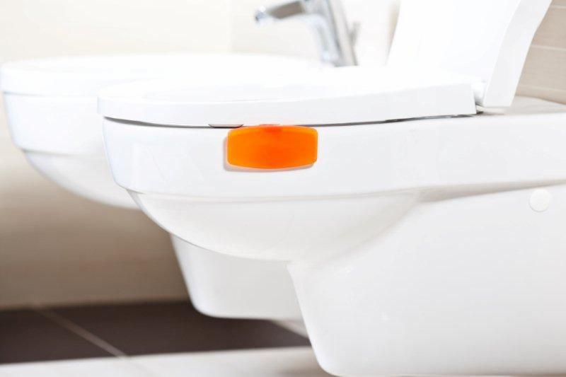 Biała toaleta z pomarańczowym odświeżaczem w kostce na krawędzi, widoczna bateria umywalkowa w tle.