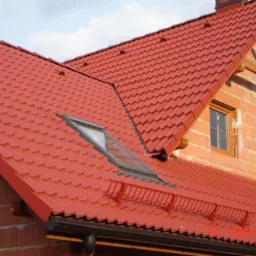 Czerwony dach z dachówką ceramiczną, okno dachowe i zabezpieczenia przeciwśniegowe na domu z cegły w trakcie budowy w słoneczny dzień.