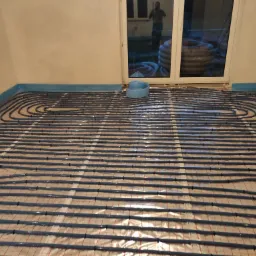 Równomiernie rozłożone rury ogrzewania podłogowego na folii izolacyjnej z widocznym fragmentem rolki rury przy oknie.