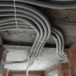 Rury instalacyjne w peszlach, przymocowane do sufitu w surowym stanie, widoczne fragmenty murowanych ścian z czerwonej cegły.