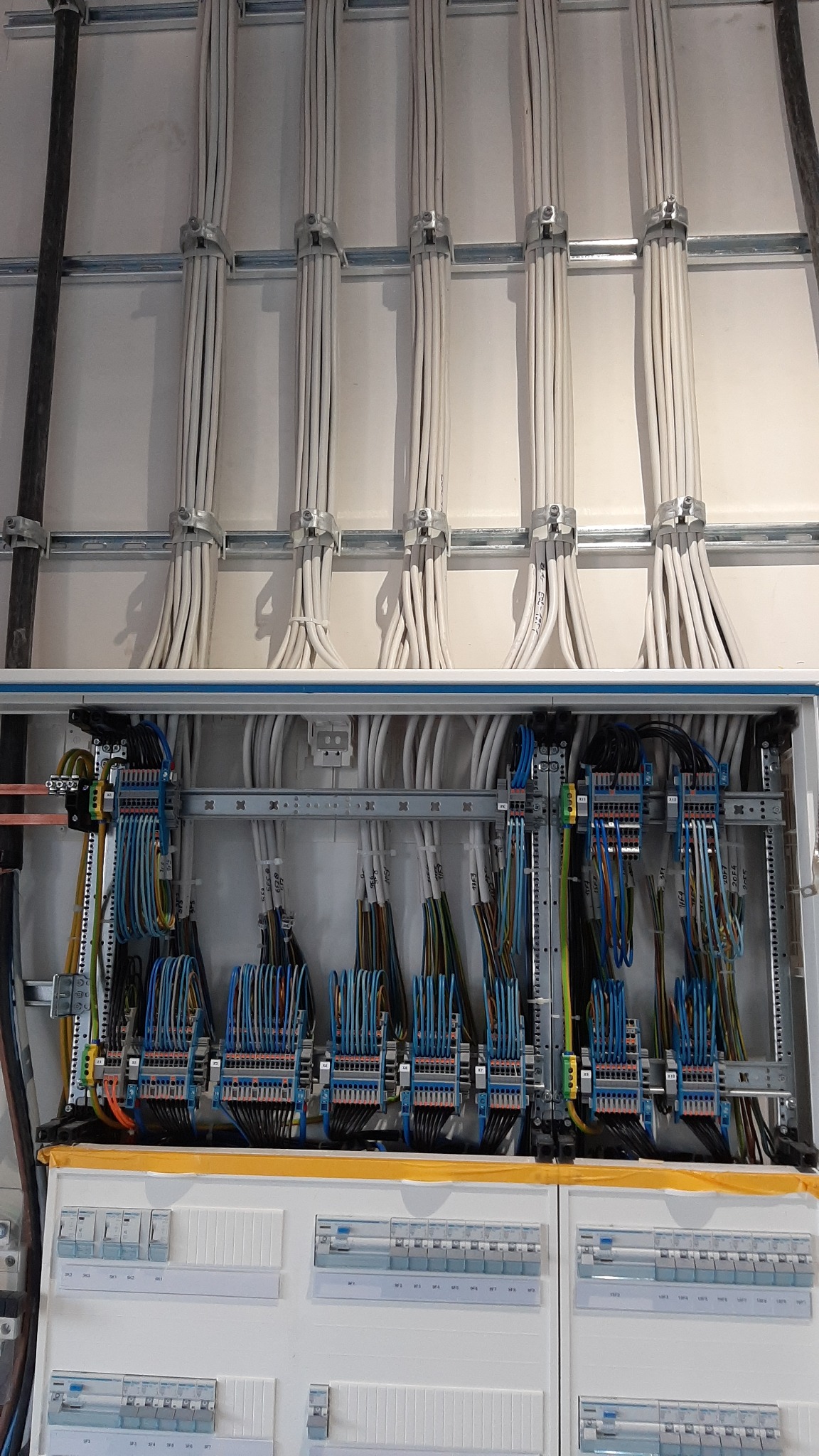 Rozdzielnica elektryczna z licznymi przewodami ułożonymi w wiązki i przymocowanymi do ściany za pomocą metalowych uchwytów, widoczne bezpieczniki i oznaczenia na przewodach.