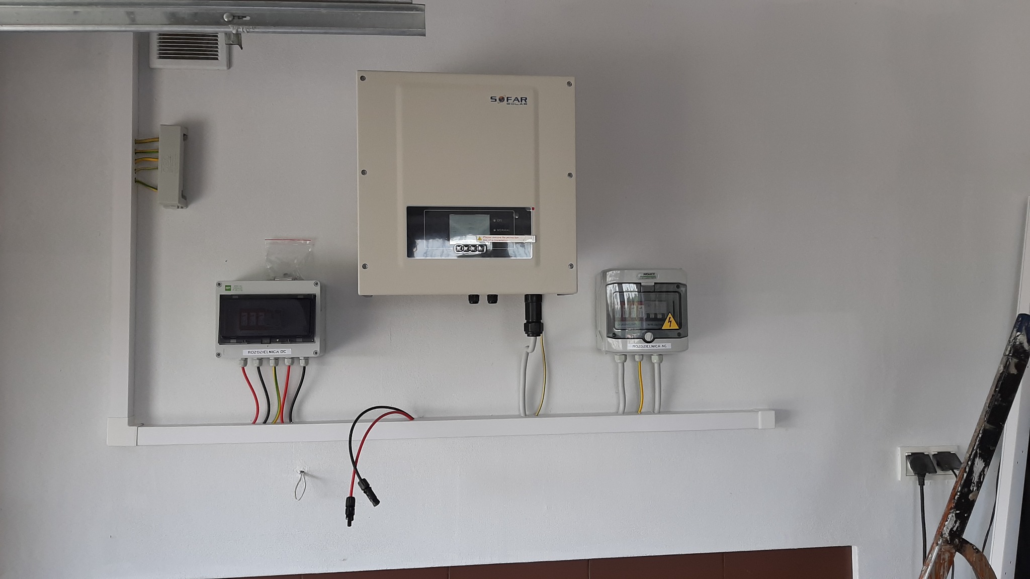 Zamontowana instalacja fotowoltaiczna wewnątrz pomieszczenia: Inwerter Sofar Solar, rozdzielnica DC z woreczkiem śrub, rozdzielnica AC, okablowanie poprowadzone wzdłuż ściany na listwie.