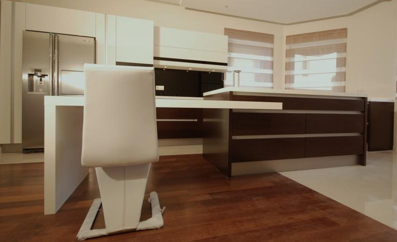 Nowoczesna kuchnia z białymi szafkami i ciemnobrązowymi szufladami, widziana zza białego krzesła z poduszkami ochronnymi na nogach, na tle drewnianej podłogi.