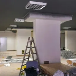 Wnętrze biura w trakcie remontu: surowy stan, widoczne kolumny, drabina oparta o ścianę, materiały budowlane i oświetlenie sufitowe.