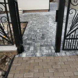 Ułożona kostka brukowa w odcieniach szarości tworząca ścieżkę w ogrodzie, widoczna brama z czarnego metalu i fragment elewacji budynku w tle.