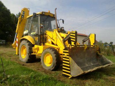 Żółta koparko-ładowarka JCB z czarno-żółtymi pasami na łyżce, stojąca na trawiastym terenie w słoneczny dzień.