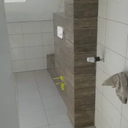 Łazienka w trakcie wykańczania: brązowe płytki imitujące drewno na zabudowie stelaża WC i obudowie półki, jasne płytki na ścianach i podłodze, widoczne żółte zaślepki na rurach, oraz zawór...