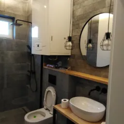 Nowoczesna łazienka z szarą płytką imitującą kamień, wisząca toaleta z zielonym odświeżaczem, umywalka nablatowa na drewnianym blacie, okrągłe lustro i czarne lampy wiszące z odsłoniętymi żarówkami.