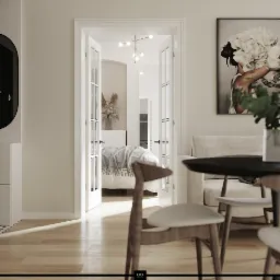 Jasne wnętrze mieszkania z widokiem na sypialnię przez przeszklone drzwi, nowoczesny żyrandol i grafika z motywem kwiatowym na ścianie, drewniane krzesła i stół na pierwszym planie.