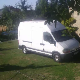 Biały bus dostawczy zaparkowany na trawniku przed budynkiem w słoneczny dzień, widok zza okna.