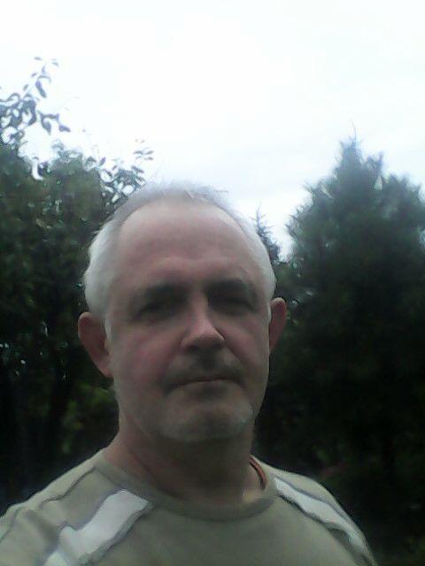 Portret mężczyzny z siwymi włosami na tle zieleni. Ujęcie z bliska, widoczna twarz i fragment ubrania. Naturalne światło, zieleń w tle.