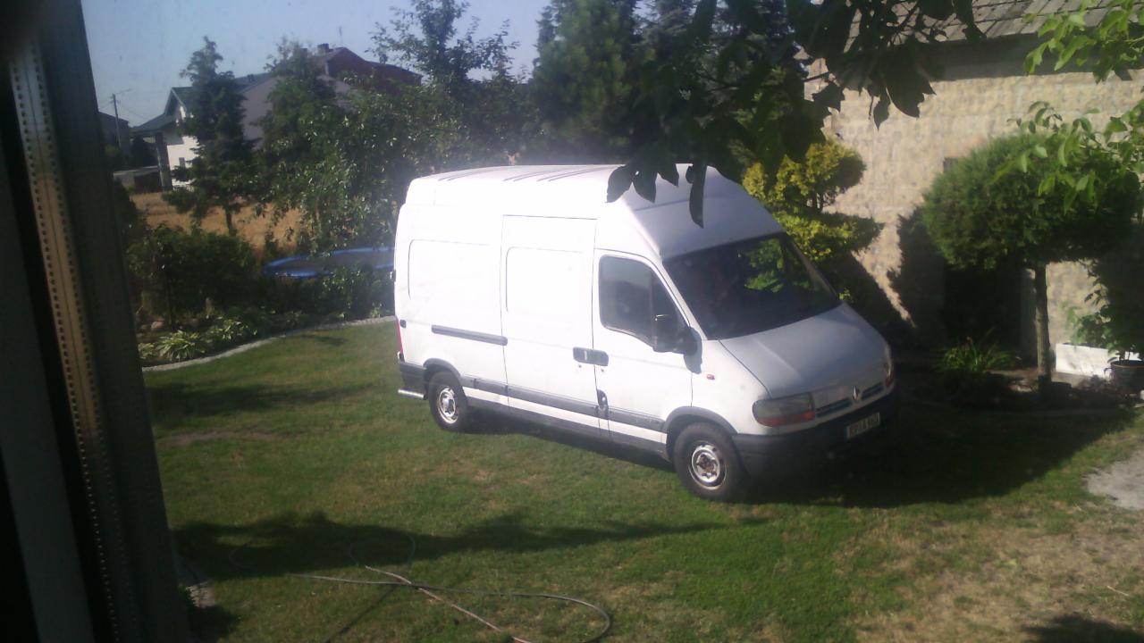 Biały bus dostawczy zaparkowany na trawniku przed budynkiem w słoneczny dzień, widok zza okna.