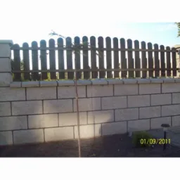 Niski murek z bloczków betonowych zwieńczony drewnianym płotkiem sztachetowym, z młodym drzewkiem posadzonym przed nim. Widoczna lampka solarna w tle.