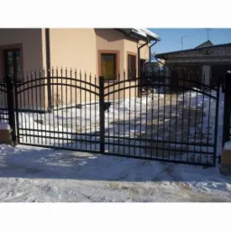 Czarna, metalowa brama skrzydłowa z dekoracyjnymi grotami i zdobieniami w zimowej scenerii przed domem z beżową elewacją i garażem.