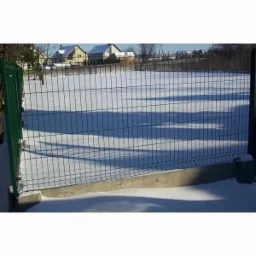 Panelowe ogrodzenie w kolorze zielonym, osadzone na betonowej podmur&oacute;wce, na tle zaśnieżonego terenu i zabudowań mieszkalnych.