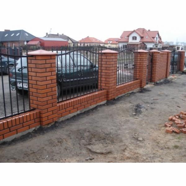 Fragment ogrodzenia z cegły klinkierowej z metalowymi przęsłami na tle zabudowy jednorodzinnej i zaparkowanego samochodu.
