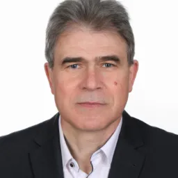 Portret mężczyzny w czarnym garniturze i białej koszuli na białym tle. Widoczna zmarszczka na czole i pieprzyk na policzku.