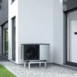 Zewnętrzna i wewnętrzna jednostka pompy ciepła powietrze-woda zamontowane przy nowoczesnym domu z białymi ścianami i szarymi elementami wykończeniowymi. Jednostka zewnętrzna stoi na metalowej...