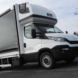 Biały samoch&oacute;d dostawczy z czarną plandeką na naczepie, zaparkowany na asfalcie, z logo firmy na nadbud&oacute;wce kabiny.