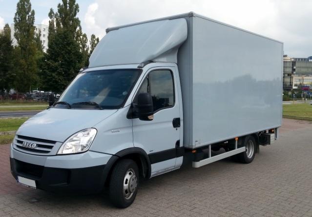 Szary bus dostawczy Iveco z wysoką zabudową kontenerową, zaparkowany na kostce brukowej, widok z boku, zieleń w tle.