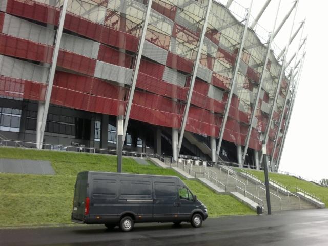Czarny bus na tle Stadionu Narodowego w Warszawie. Auto zaparkowane na mokrym asfalcie, widok z boku, pochmurna pogoda.