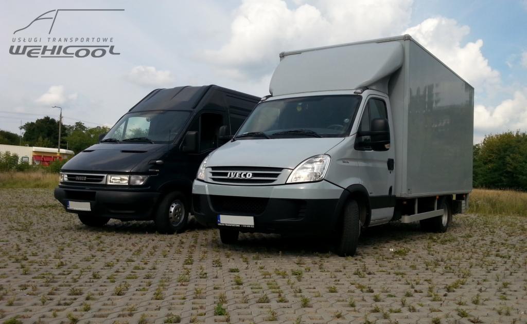 Dwa busy Iveco: czarny furgon i srebrny z zabudową kontenerową, stojące na placu z betonowej kostki, pod błękitnym niebem z chmurami. Widoczne logo firmy transportowej.