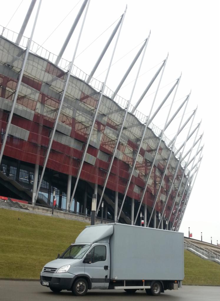 Szary bus dostawczy na tle Stadionu Narodowego w Warszawie, pochmurny dzień. Perspektywa z dołu, ukazująca charakterystyczną architekturę stadionu.