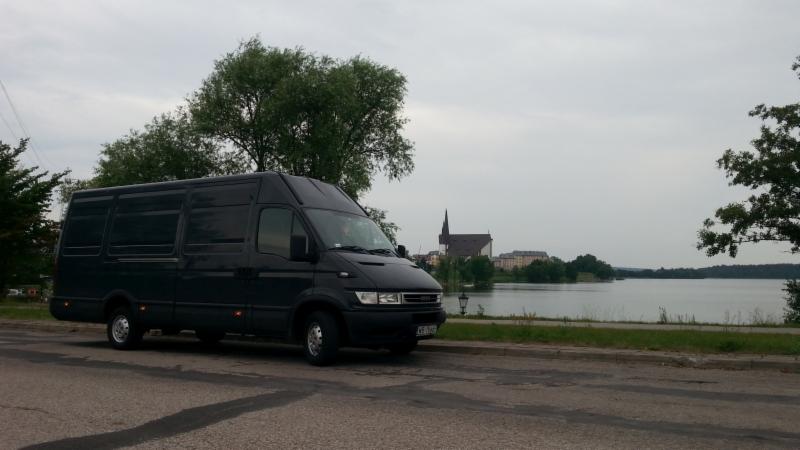 Czarny bus na tle jeziora i kościoła. Pojazd zaparkowany na poboczu, widoczne drzewa i pochmurne niebo. Bus marki Iveco.