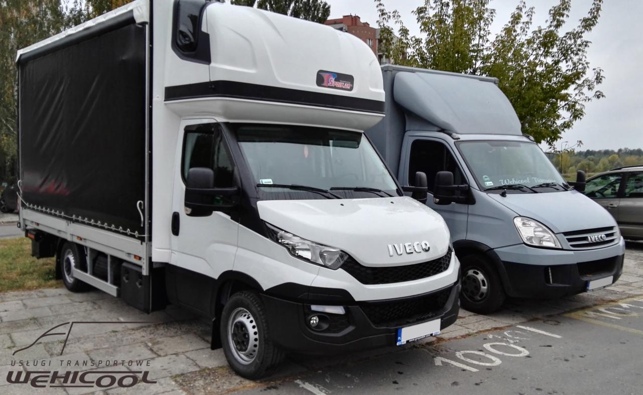 Dwa busy Iveco, biały z czarną plandeką i szary, zaparkowane na kostce brukowej. Na białym busie widoczne logo 'Sportrar'.