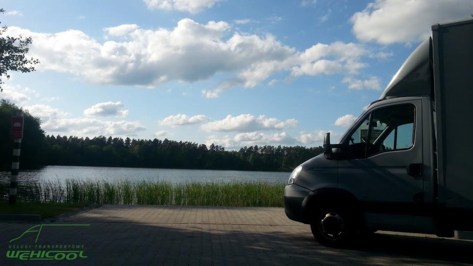 Szary bus dostawczy na tle jeziora i lasu, błękitne niebo z chmurami. Widoczne logo firmy transportowej. Ujęcie z dołu, podkreślające przestrzeń.