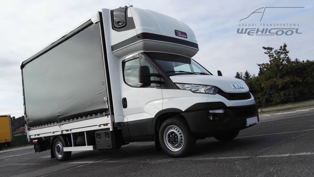 Biały samochód dostawczy z czarną plandeką na naczepie, zaparkowany na asfalcie, z logo firmy na nadbudówce kabiny.