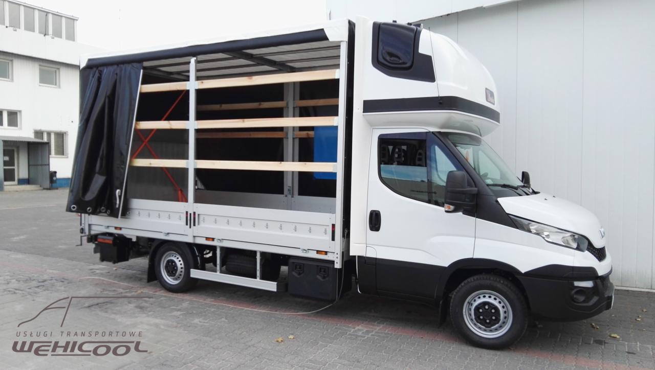 Biały samochód dostawczy z otwartą przestrzenią ładunkową, zabezpieczoną czarną plandeką i drewnianymi belkami, widoczny z boku na tle szarej kostki brukowej i białego budynku.