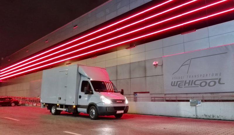Szary bus dostawczy Iveco z zabudową kontenerową na tle budynku z neonowym oświetleniem i logo firmy transportowej WEHICOOL. Nocne ujęcie z dołu.