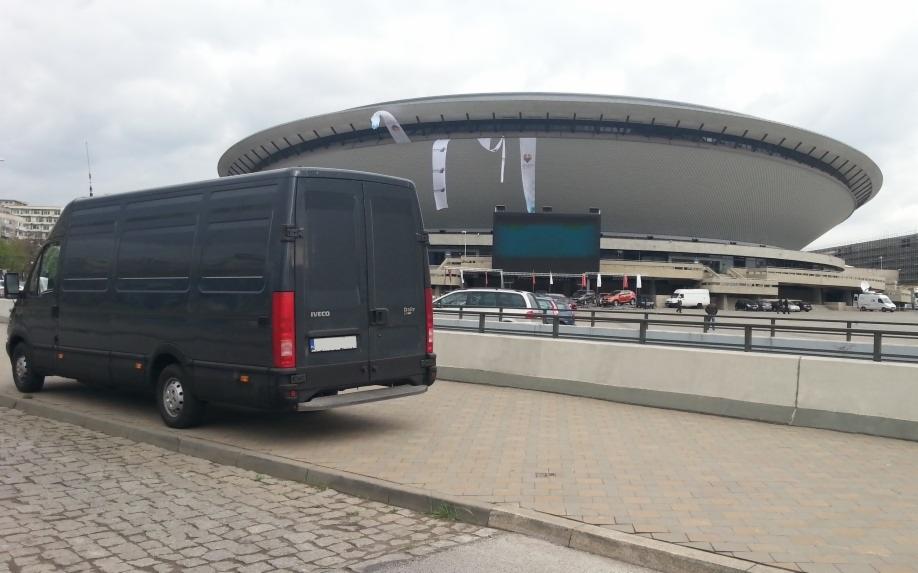 Czarny bus Iveco parkuje na tle Spodka w Katowicach. Widok z tyłu na pojazd, z widocznymi światłami i tablicą rejestracyjną. Pochmurne niebo.