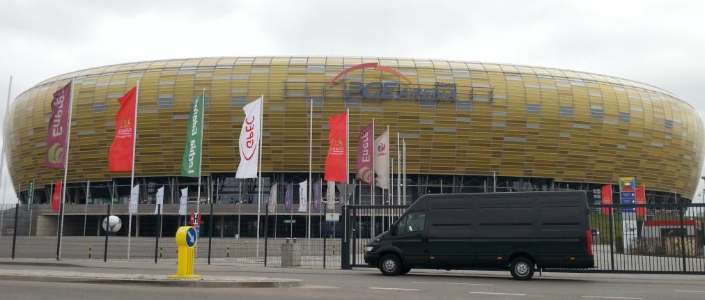 Czarny bus na tle PGE Areny w Gdańsku, z flagami reklamowymi. Pojazd zaparkowany przed bramą wjazdową. Szary dzień.