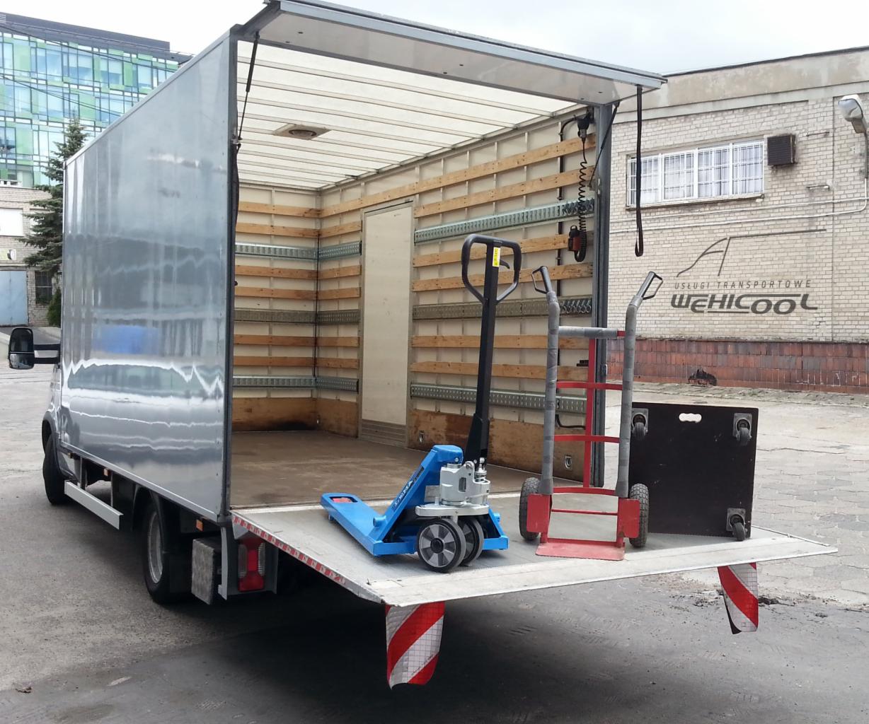 Półotwarty bus transportowy z rampą załadowczą, wewnątrz wózki paletowe i transportowe. Widoczny budynek z logo firmy transportowej.