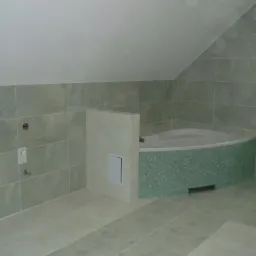 Wykończona łazienka na poddaszu z wanną obłożoną mozaiką i jasnymi płytkami na ścianach i podłodze. Widoczne przyłącza wodne i wentylacja.