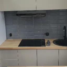 Nowoczesna kuchnia z szarymi kafelkami, czarnym zlewem i płytą indukcyjną, drewnianym blatem oraz beżowymi szafkami, widoczny fragment osoby montującej meble.