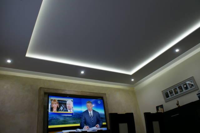 Sufit podwieszany z oświetleniem LED w nowoczesnym salonie. Widoczne lampy punktowe i telewizor. Jasne ściany w tle.