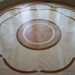 Dekoracyjna podłoga z parkietu ułożonego we wzór rozety z centralnym okręgiem i motywem kwiatowym, wykonana z różnych odcieni drewna.