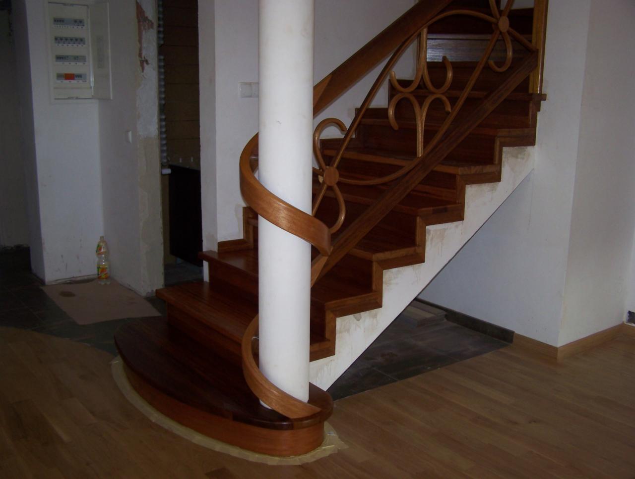 Drewniane schody z balustradą o dekoracyjnych wycięciach, owijające się wokół białej kolumny, z widocznym fragmentem podłogi z paneli.