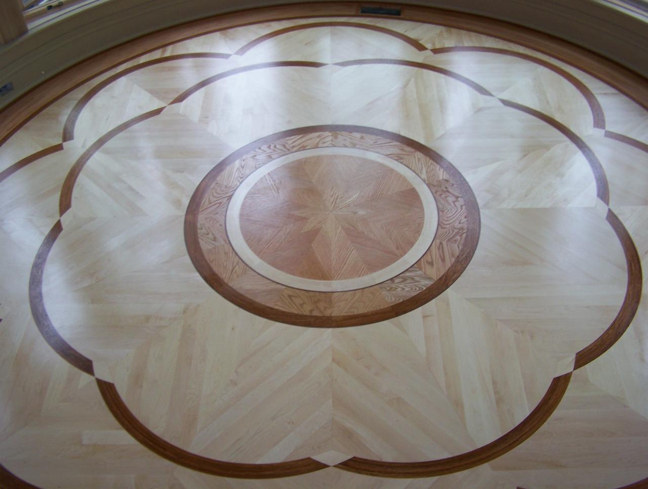 Dekoracyjna podłoga z parkietu ułożonego we wzór rozety z centralnym okręgiem i motywem kwiatowym, wykonana z różnych odcieni drewna.