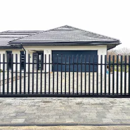 Nowoczesna brama wjazdowa z pionowych, czarnych profili stalowych, osadzona na szarych, betonowych słupkach, Bystra. W tle widoczny dom z garażem i szarą dachówką.