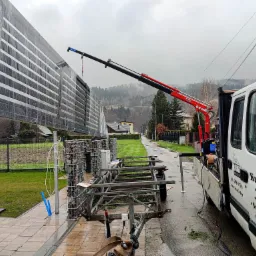 Montaż ogrodzenia panelowego z użyciem dźwigu samochodowego, panel w folii ochronnej, widoczny fragment zabudowy gabionowej i przyczepa transportowa, pochmurny dzień.