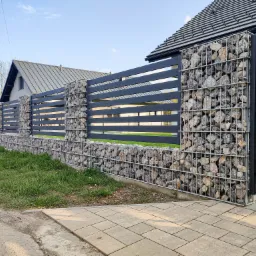 Nowoczesne ogrodzenie z poziomych, grafitowych paneli metalowych i gabionowych słupków wypełnionych kamieniami, na tle domów z szarymi dachami.