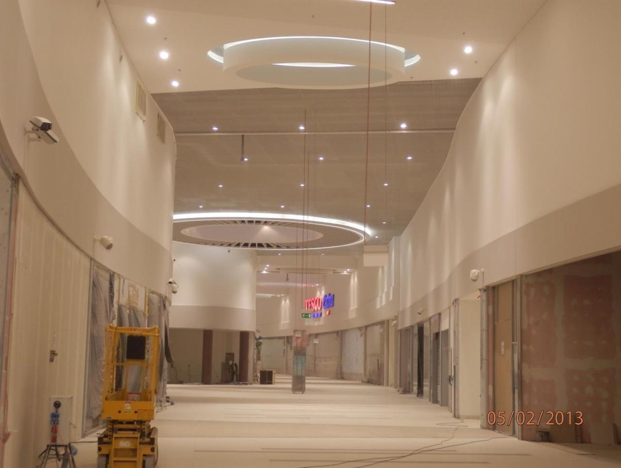 Montaż podwieszanego sufitu z oświetleniem w przestrzeni handlowej, widoczny podnośnik i niedokończone ściany.